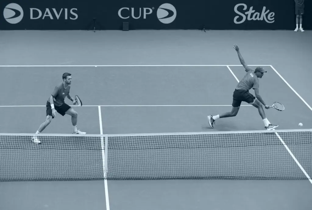 שני שחקני טניס בזוגות בגביע דייויס, האחד תוקף והשני ממתין, עם לוגו וטורניר פוקר וקזינו ברקע. stakeil -